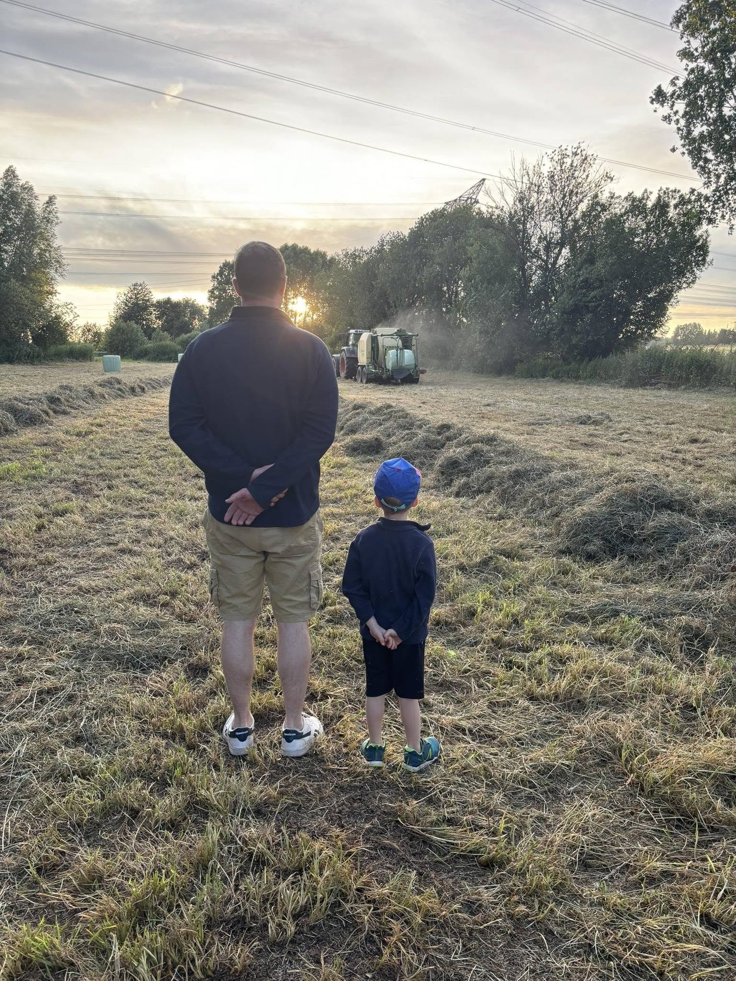 Un père et son fils, de dos, regardent le pressage du foin au coucher du soleil